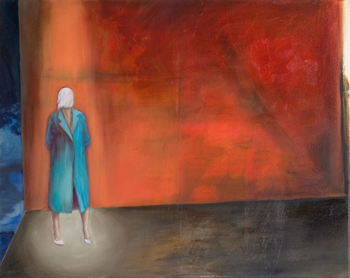 eine Frau im blauen Mantel und weißen Tuch vorm Gesicht steht vor orangeroter Wand, ein Spot auf ihr